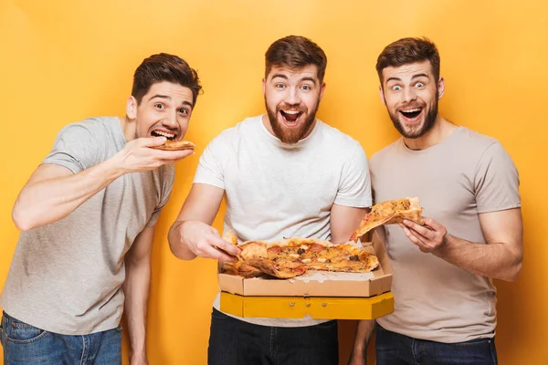 Three young happy men eating big pizza isolated over yellow background