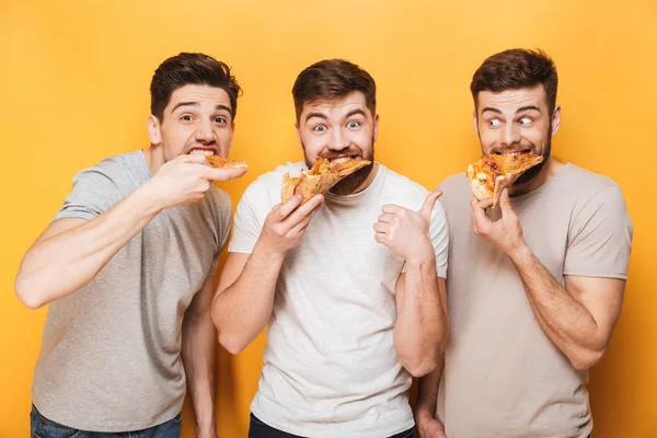 Three young happy men eating pizza isolated over yellow background