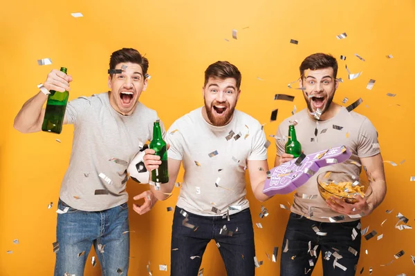 Three young joyful men football fans celebrating with beer and snacks isolated over yellow background