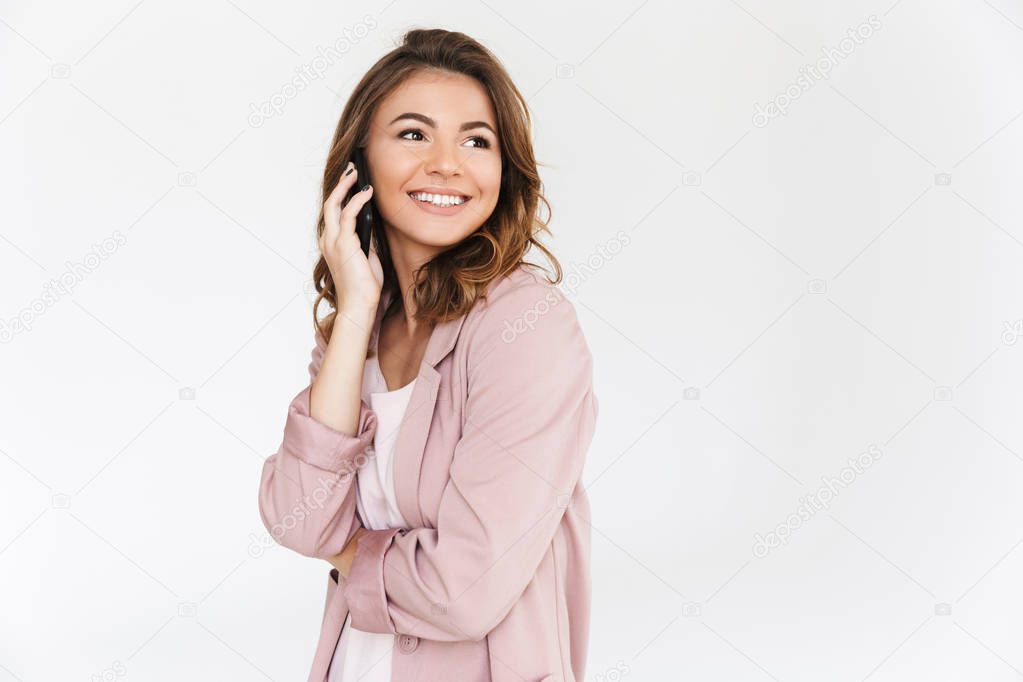 Photo of young cute lady standing isolated over white wall background talking by mobile phone.