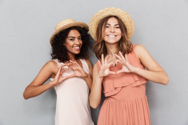 Yaz elbise gri arka plan üzerinde izole silah ile kalp hareketi gösteren iki genç kadın gülümseyerek portresi giymiş