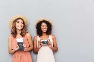 Pasaport ve bilet ve fotoğraf makinesi gri arka plan üzerinde tutarak yaz elbise giymiş iki genç kadın gülümseyerek portresi