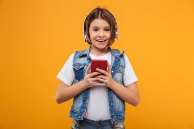 Sarı arka plan üzerinde cep telefonu tutarken kulaklık ile müzik dinlemek bir neşeli küçük kız öğrenci portresi