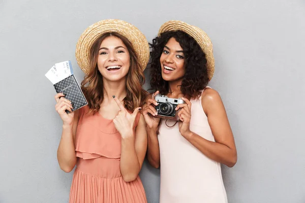 Pasaport ve bilet ve fotoğraf makinesi gri arka plan üzerinde tutarak yaz elbise giymiş iki neşeli genç kadın portresi