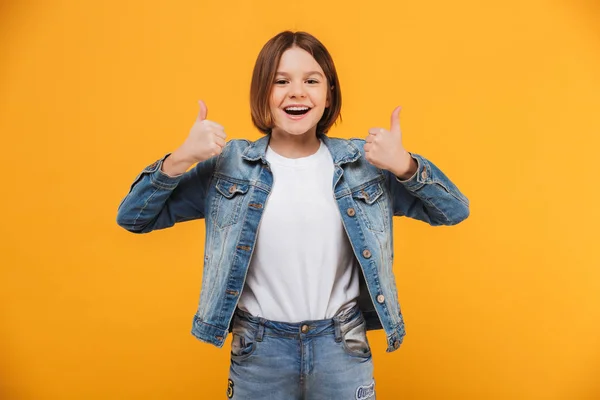Portrait of an excited little schoolgirl showing thumbs up gesture over yellow background