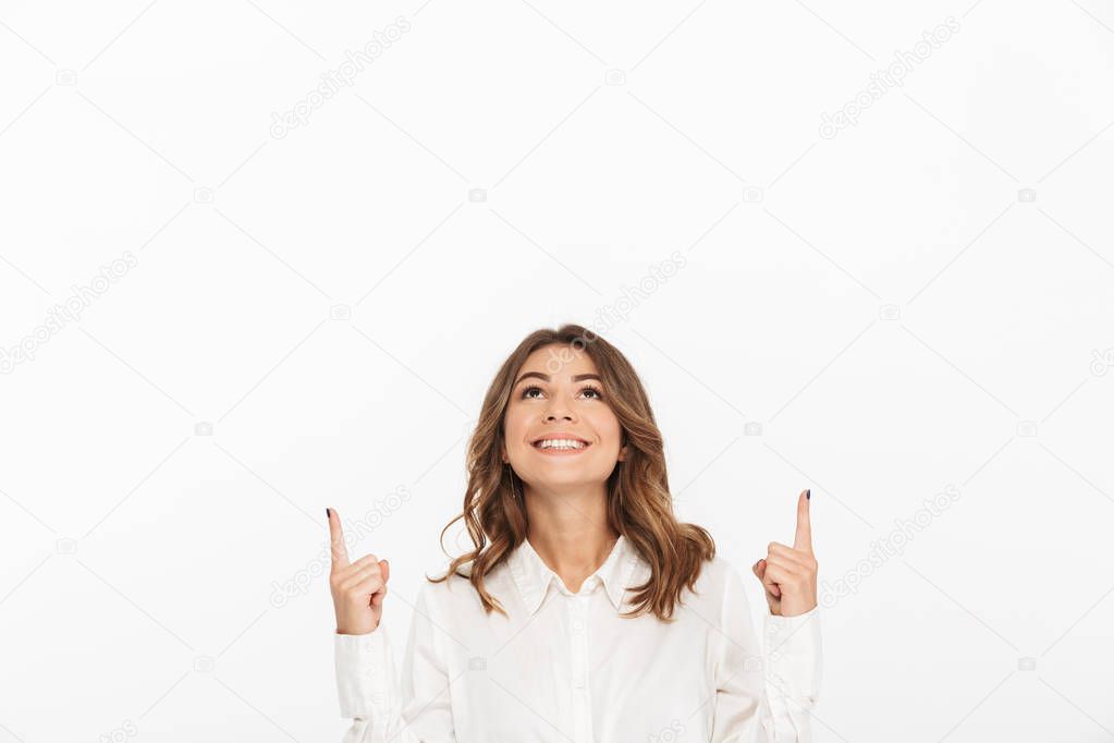 Portrait of a smiling young business woman pointing fingers up at copy space isolated over white background