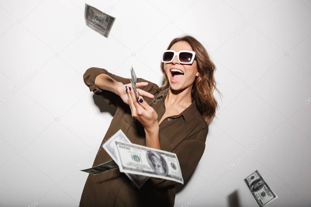 Portrait of a happy young woman in sunglasses throwing out money banknotes isolated over white background