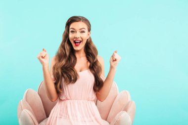 Pembe koltukta oturan ve üzerinde mavi arka plan izole yumruk kenetlenmesi ile sürpriz içinde bağırmaya sürpriz kadın giyiyor elbise görüntü