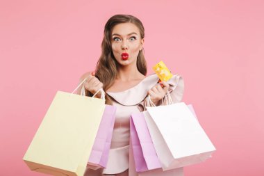 Uzun kahverengi saçlı renkli alışveriş torbaları ve kredi kartı üzerinde pembe arka plan izole elinde tutarken gülümseyen içinde memnun büyüleyici kadın 20'li