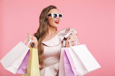 Elbise ve renkli alışveriş torbaları üzerinde pembe arka plan izole gülümseme ile tutarak şık güneş gözlüğü takmış uzun kahverengi saçlı memnun büyüleyici kadın