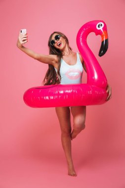 Genç ve güzel bir kadın portresi tam uzunlukta mayo ile şişme flamingo üzerinde pembe arka plan izole bir selfie alırken poz giyinmiş