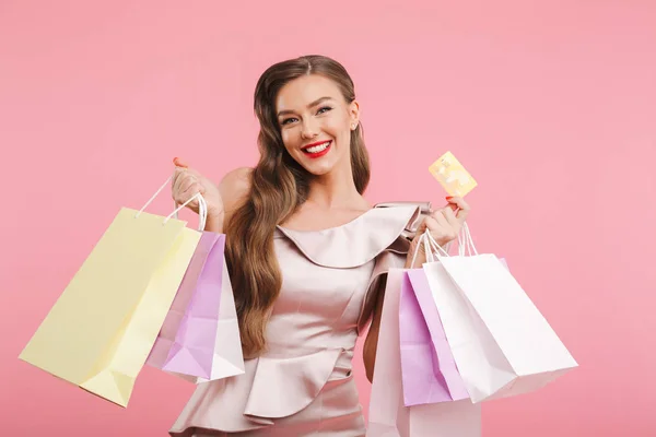 时髦的女人20S 穿着长棕色的头发微笑着 而持有不同的购物包和信用卡在手被孤立的粉红色背景 — 图库照片
