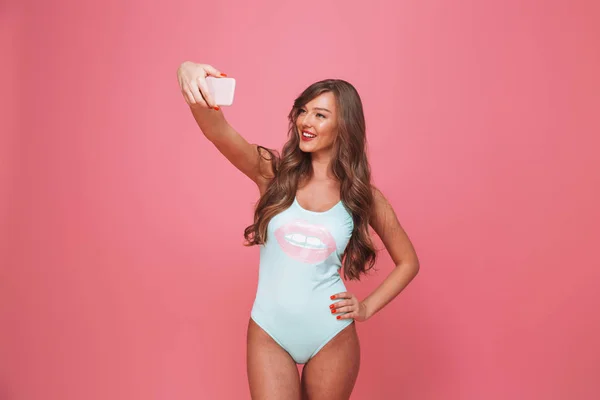 穿着泳装的微笑的年轻妇女的画像采取自拍用移动电话隔绝了在粉红色背景下 — 图库照片