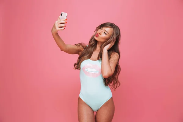 一个性感的年轻女子的肖像穿着泳装采取自拍与手机在粉红色背景下隔离 — 图库照片