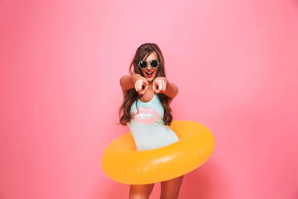 一个兴奋的年轻女子的肖像 穿着泳装摆着充气戒指和指向在粉红色背景隔离相机 — 图库照片
