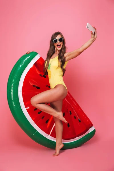 幸せな若い女性の完全な長さの肖像画は膨脹可能なスイカと立っているとピンク背景に分離された Selfie を撮影中ポーズ水着に身を包んだ — ストック写真