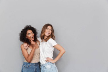 Portrait of two happy young women standing together and looking away at copy space isolated over gray background clipart