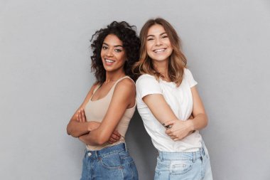 Portrait of two cheerful young women standing together and looking at camera isolated over gray background clipart