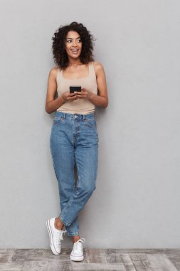 Cep telefonunuzun ve gri arka plan üzerinde gülümseyen genç Afrikalı kadın portresi tam uzunlukta