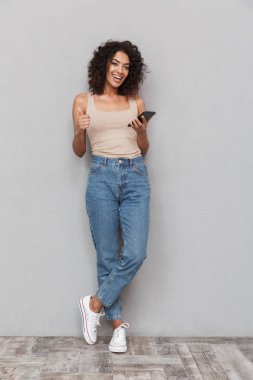 Hareket eden telefon tutarak ve gri arka plan üzerinde başparmak gösterilmesini bir gülümseyen genç Afrikalı kadın portresi tam uzunlukta