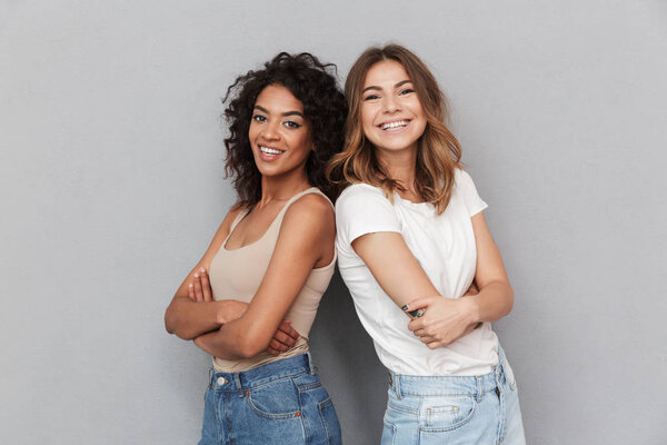 Portrait of two cheerful young women standing together and looking at camera isolated over gray background