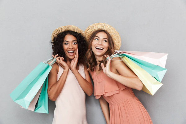 Portrait of two happy young women dressed in summer clothes holding shopping bags isolated over gray background