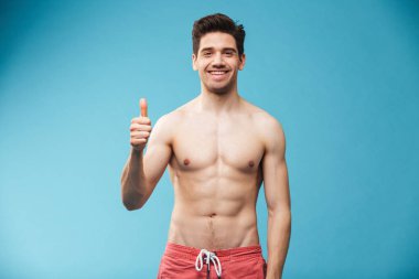 Mavi arka plan üzerinde gösteren başparmak yüzme gülümseyen bir genç gömleksiz adam portresi şort