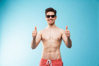 Yüzme şort ve mavi arka plan üzerinde başparmak gösterilmesini güneş gözlüğü içinde mutlu bir genç gömleksiz adam portresi