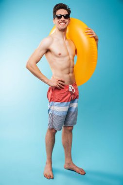 Tam boy şişme halka içinde güneş gözlüğü mavi arka plan üzerinde poz ile neşeli bir genç gömleksiz adam portresi