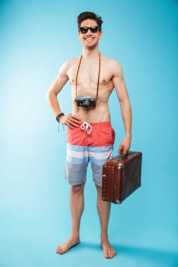 Yüzme neşeli bir genç gömleksiz adam portresi tam uzunlukta taşıma fotoğraf makinesi ve bavul mavi arka plan üzerinde şort