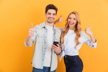 Neşeli çift erkek ve kadın gülümseyerek ve başparmak kadar gösteren izole sarı arka plan üzerinde siyah cep telefonu tutarken fotoğraf