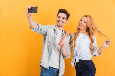 İki mutlu insanlar erkek ve kadın selfie smartphone barış sarı arka plan üzerinde izole işareti gösterilen süre fotoğrafta portre