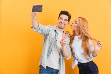 Güzel çift sarı arka plan üzerinde izole yanağında adamı öperken kadın sırasında cep telefonuyla selfie fotoğrafta portresi