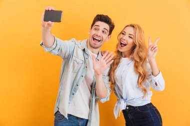 Mutlu bir adam ve kadın selfie cep telefonu ile izole sarı arka plan üzerinde parmak kameraya el hareketi sırasında fotoğrafta portre