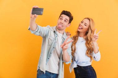 İki komik insanlar erkek ve kadın selfie smartphone üzerinde zafer işareti izole sarı arka plan üzerinde gösterilen süre fotoğrafta portre