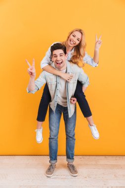 Eğleniyor ve zafer işareti adam gülümseyen kadın sarı arka plan üzerinde izole piggybacking yaparken gösterilen çift tam boy fotoğrafı