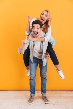 Eğleniyor ve boşaltmak izole sarı arka plan üzerinde neşeli kadın piggybacking adam, parmakları işaret çift tam boy görüntü