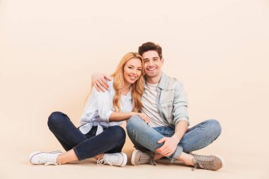 Görüntü güzel ve şirin çift denim içinde bej renkli arka plan üzerinde giyim gülümseyen ve bacaklar ile katta oturan geçti iken birlikte sarılma izole