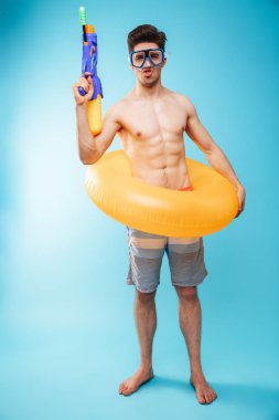 Komik bir genç gömleksiz adam yüzmek gözlük ve şişme yüzük eğleniyor su tabancası ile mavi arka plan üzerinde tam uzunlukta portresi