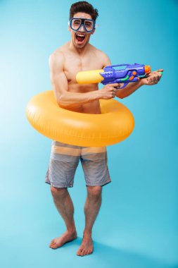 Genç, neşeli bir gömleksiz adam yüzmek gözlük ve şişme yüzük eğleniyor su tabancası ile mavi arka plan üzerinde tam uzunlukta portresi