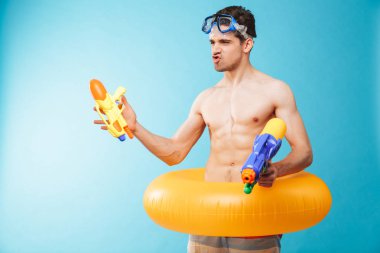 Yüzmek gözlük ve şişme yüzük eğleniyor su silahlar ile mavi arka plan üzerinde heyecanlı genç gömleksiz adam portresi