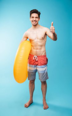 Tam boy şişme halka başparmak mavi arka plan üzerinde göstermek yukarıya ile mutlu bir genç gömleksiz adam portresi