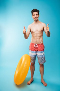 Tam boy şişme halka başparmak mavi arka plan üzerinde göstermek yukarıya ile neşeli bir genç gömleksiz adam portresi