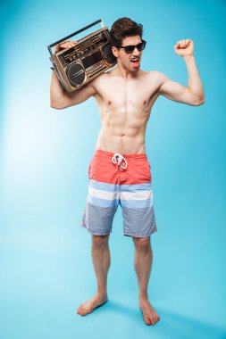 Tam uzunlukta portre Eğer kayıt cihazı tutarak ve mavi arka plan üzerinde dans yüzme şortu giymiş bir neşeli gömleksiz adam