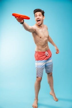 Tam uzunlukta portre Eğer üzerinde mavi arka plan ile frizbi oynayan shorts yüzme içinde neşeli bir gömleksiz adam