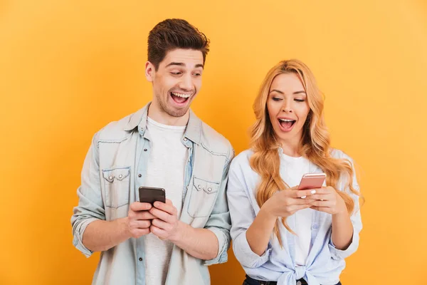 面白い若いカップル男女お互いをピークする黄色背景に分離の手で保持する携帯電話の画像 — ストック写真