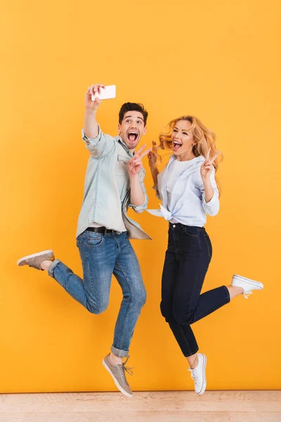 Full length photo of happy man and woman taking selfie photo on mobile phone while jumping and showing victory sign isolated over yellow background