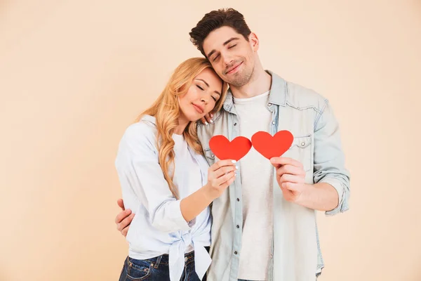 Portrait of joyful romantic people man and woman in basic clothing holding red paper hearts on sticks and hugging with closed eyes isolated over beige background