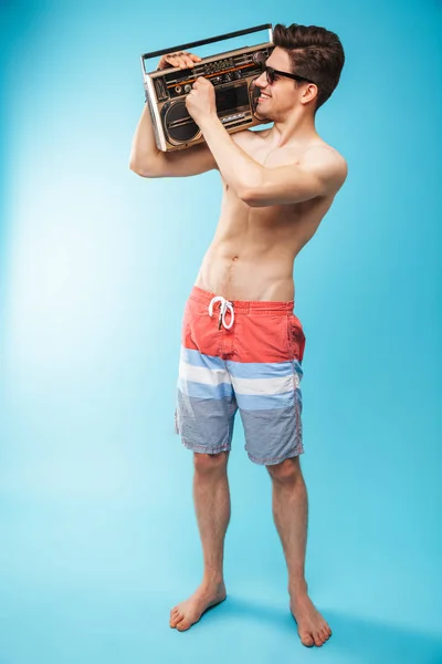 完全な長さの肖像画場合テープ レコーダーを押し 青い背景に楽しい水泳パンツでうれしそうな上半身裸の男 — ストック写真