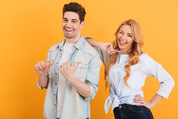 Photo of two young people in basic clothing smiling and pointing finger aside at copyspace isolated over yellow background